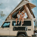 Two people sitting on the roof of a beige camper van with an open roof against a blue sky.