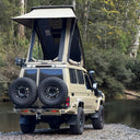 Beige off-road vehicle with a rooftop tent parked near a body of water with trees in the background.