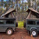 Two green off-road vehicles with rooftop tents parked in a forest.