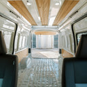 The interior rear view of a white van with wood-look ceiling panels, a textured metal floor, and dark blue-gray seats, with the back door open showing a bright outdoor patio beyond.