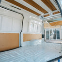 The interior side wall and cargo area of a white van with wood-look ceiling panels and unfinished tan paneling on the side walls.