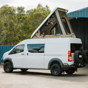 A white van with a large beige and black pop-up roof tent mounted above it, parked outdoors in front of a blue metal building with trees in the background.