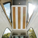 A white van interior ceiling with three vertical wood-grain panels mounted on a white frame, surrounded by windows showing outdoor scenery and bright sunlight.