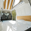 The interior of a white van with the rear doors open, showing a spacious cargo bed with a textured floor, light wood ceiling panels, and a wooden back wall panel.